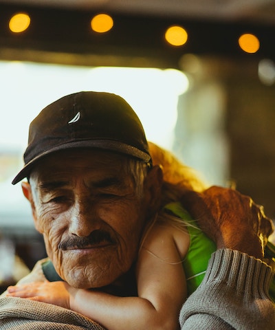 A grandfather smiles slightly while hugging a small child resting on his shoulder
